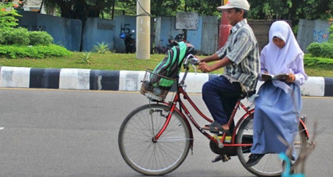 Seorang pelajar sedang membaca buku saat dibonceng orang tuanya naik sepeda ke sekolah. Foto: ilustrasi dokumen JPNN